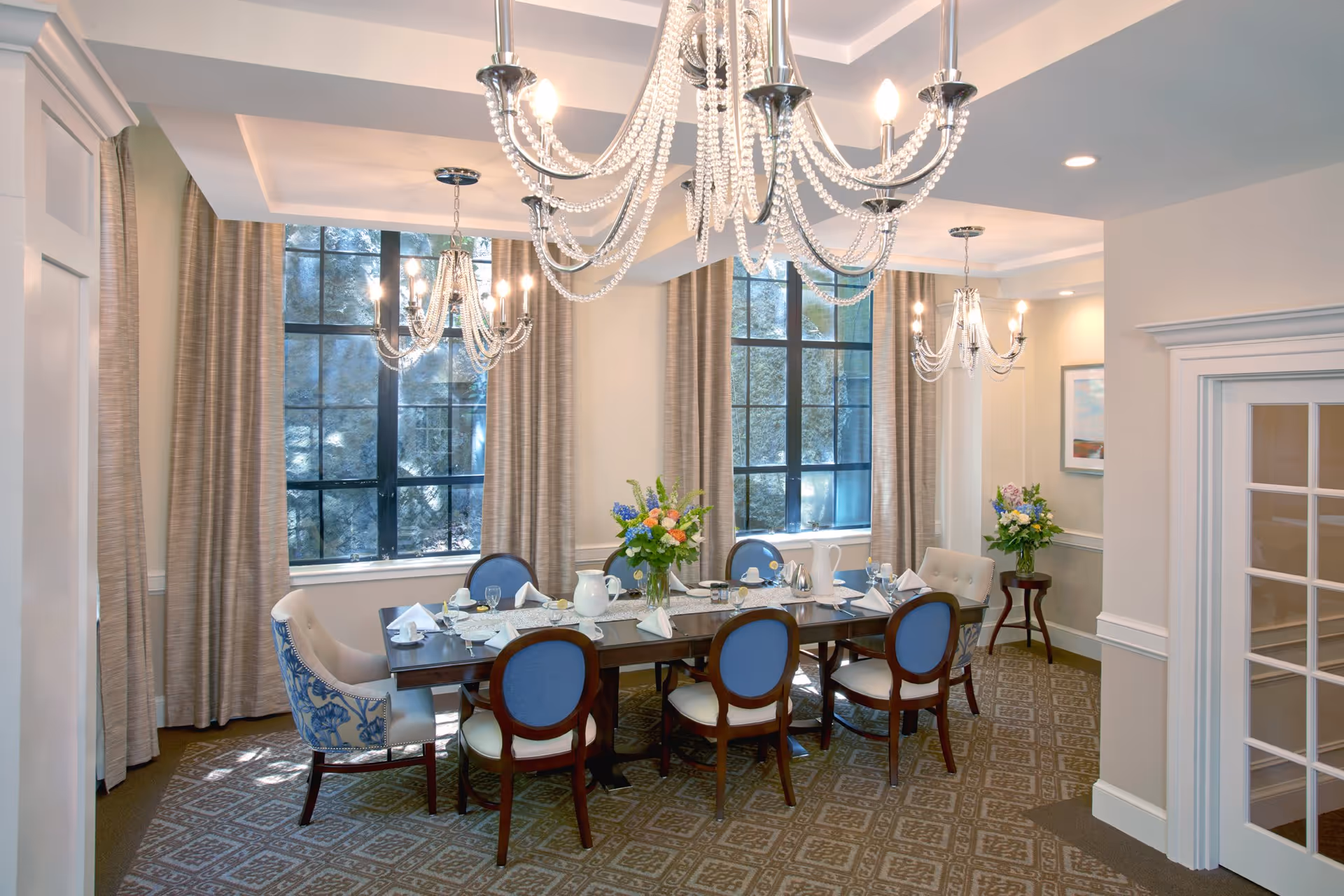 Elegant dining room with a long wooden table set for a meal, surrounded by eight chairs with blue and white upholstery. The room features large windows with beige curtains, three ornate chandeliers hanging from the ceiling, and floral centerpieces on the table and a small side table. The walls are painted in a light color with white trim and molding.