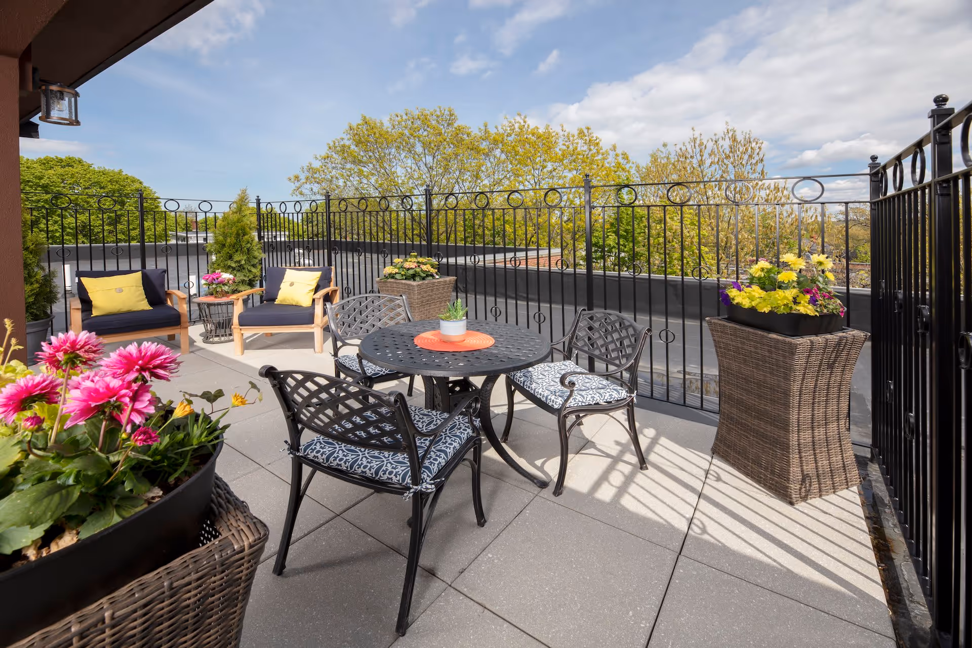 Outdoor patio area with a black metal table and four chairs with patterned cushions. Two wooden armchairs with navy cushions and yellow pillows are placed near the corner. Several planters with colorful flowers and greenery are arranged around the space. The patio is enclosed by a black metal fence, and trees with green leaves are visible in the background under a partly cloudy sky.