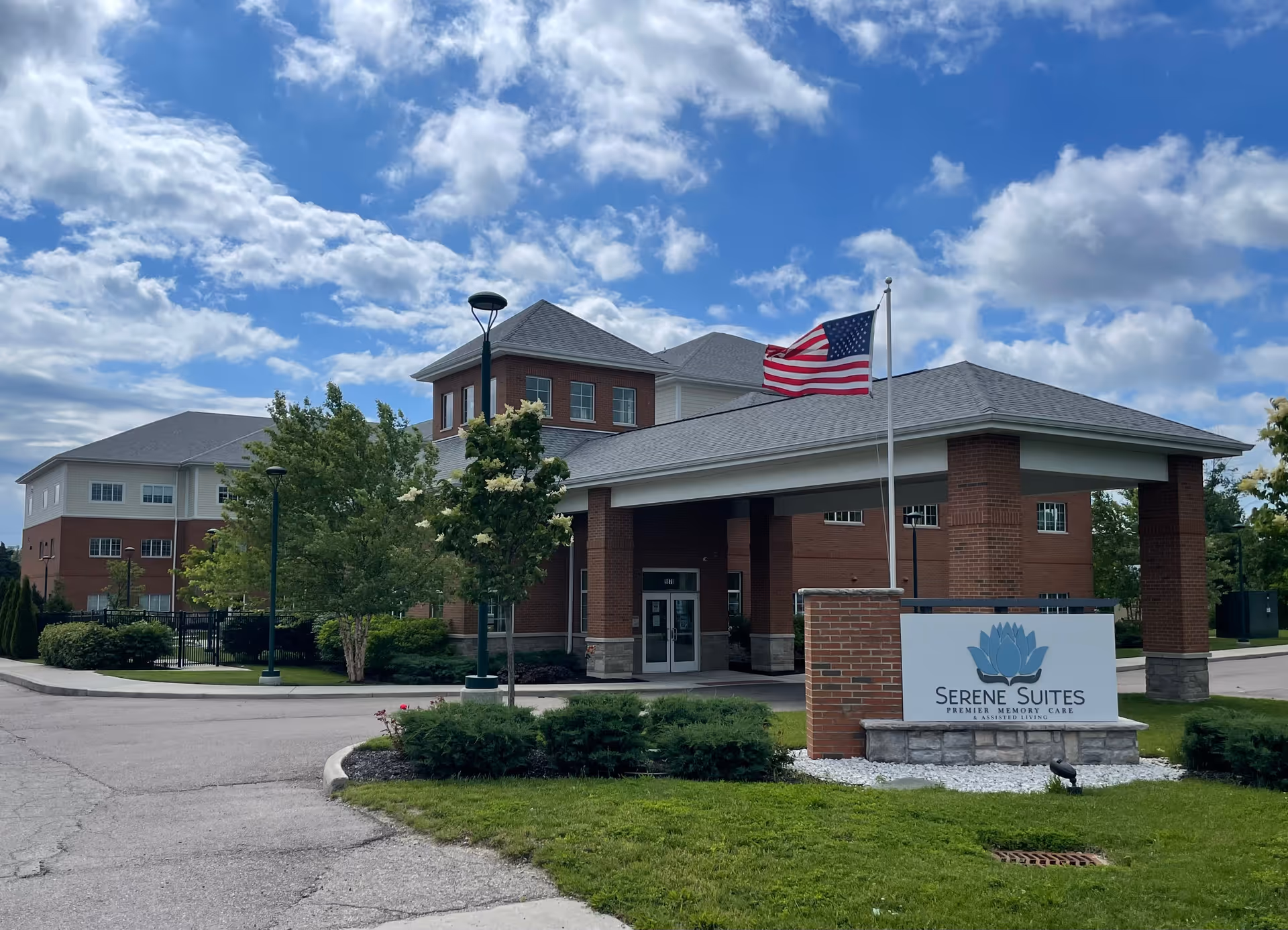 Exterior view of Serene Suites, a multi-story brick and siding building with a covered entrance. An American flag is flying on a flagpole near the entrance. The sky is partly cloudy and there are trees and shrubs around the building.