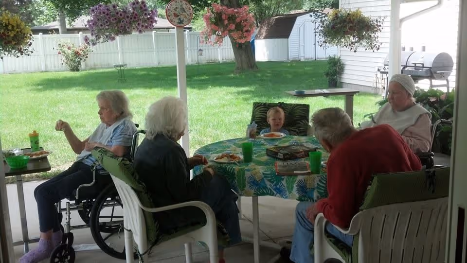 A group of elderly people and a young child sitting around a table outdoors under a covered patio, enjoying a meal together. The table is covered with a colorful tablecloth and there are hanging flower baskets and a green lawn with a white fence in the background.