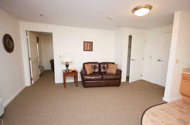 Interior view of a living room area in a senior living facility with a brown leather loveseat, two patterned throw pillows, a wooden side table with a lamp and decorative items, beige carpet, white walls, and multiple doorways leading to other rooms.