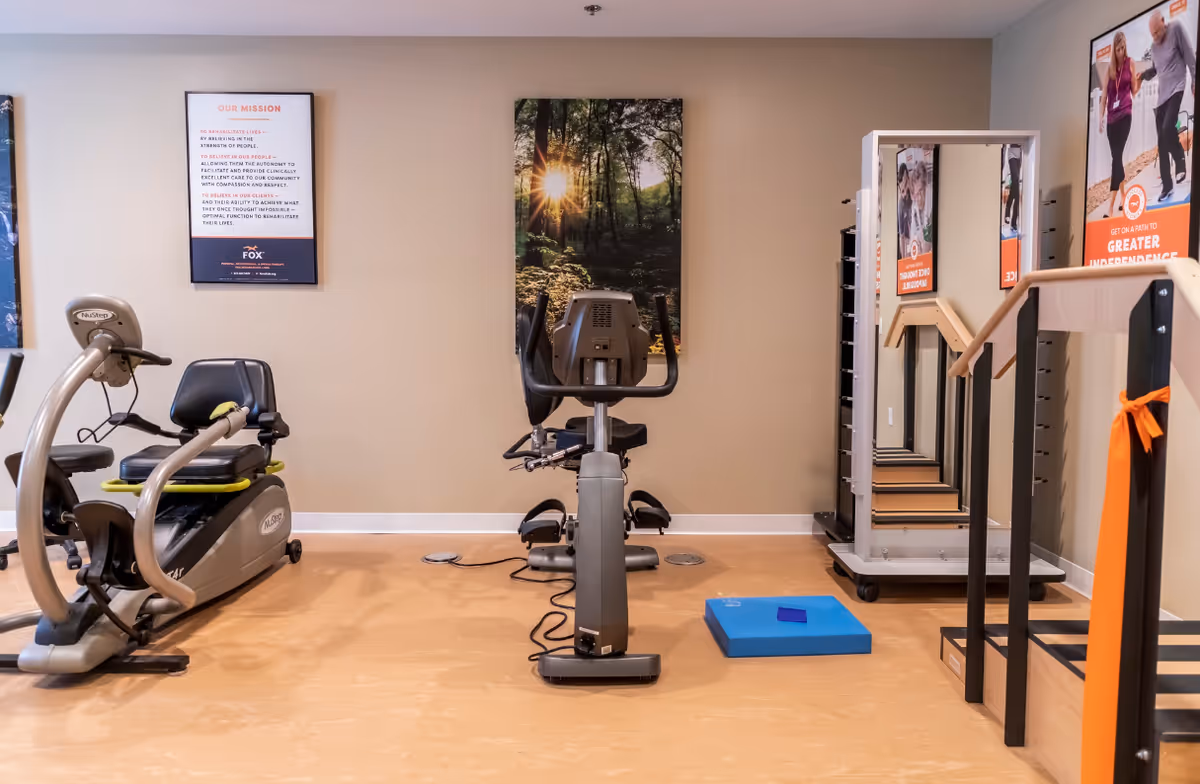 A fitness room with exercise equipment including a recumbent bike and an upright stationary bike. There is a set of stairs with handrails on the right side, a blue exercise mat on the floor, and a large mirror with a rack for weights. The walls are beige with framed posters and a nature-themed picture.
