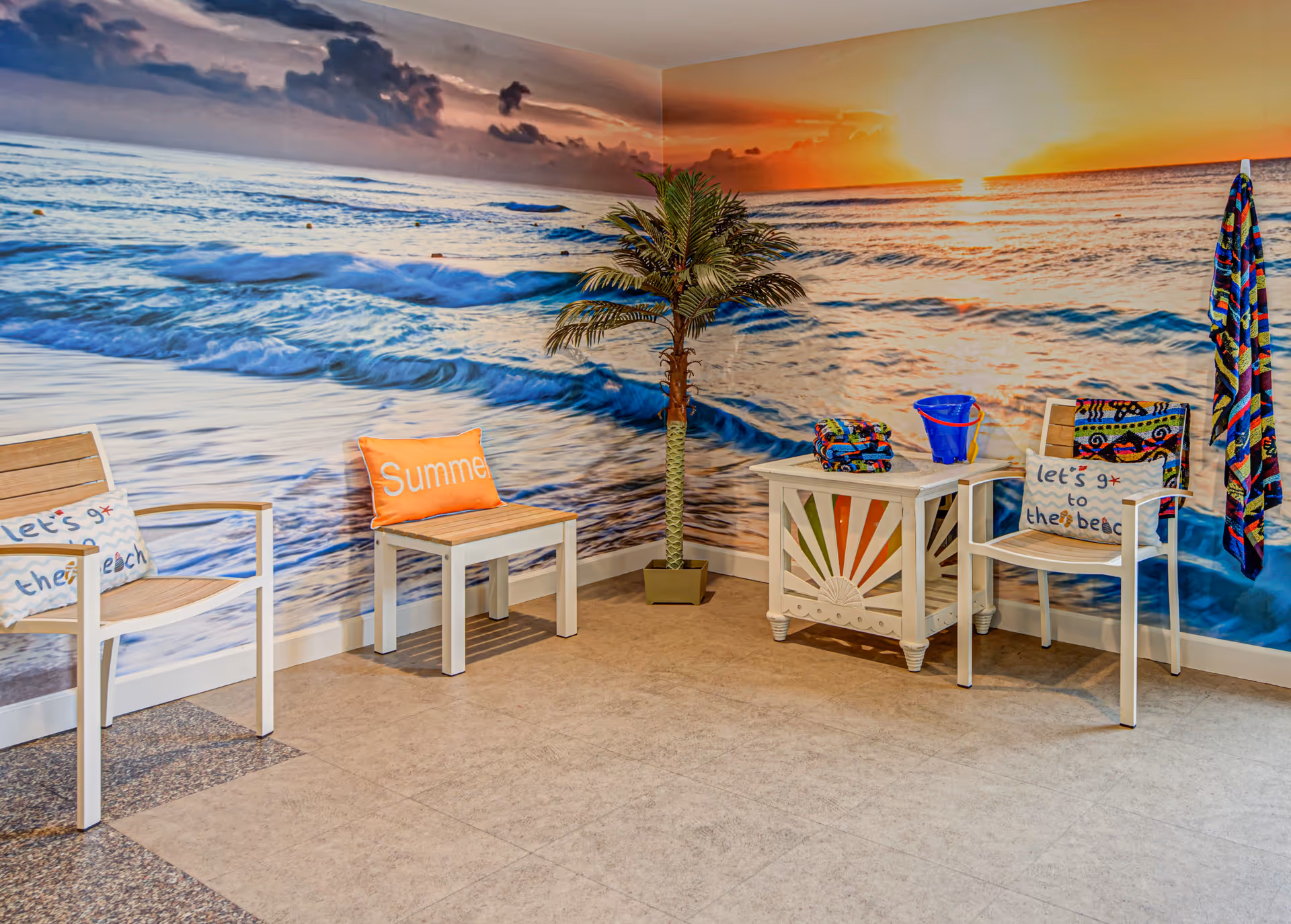 Indoor seating area with two white chairs and a small white table. The chairs have beach-themed pillows, one says 'let's go to the beach' and the other says 'Summer'. A colorful towel hangs on the wall, and a potted artificial palm tree stands in the corner. The walls are decorated with a large mural of a beach scene at sunset with waves and clouds.