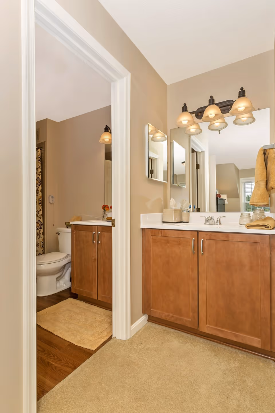 View of a bathroom area with a wooden vanity cabinet topped with a white countertop and a large mirror above it. The vanity has a silver faucet and several items on the counter including a tissue box and jars. To the left, there is an open doorway leading to a toilet area with a patterned shower curtain and a beige bath mat on the wooden floor. The walls are painted beige and the lighting fixture above the mirror has five lamps with frosted glass shades.