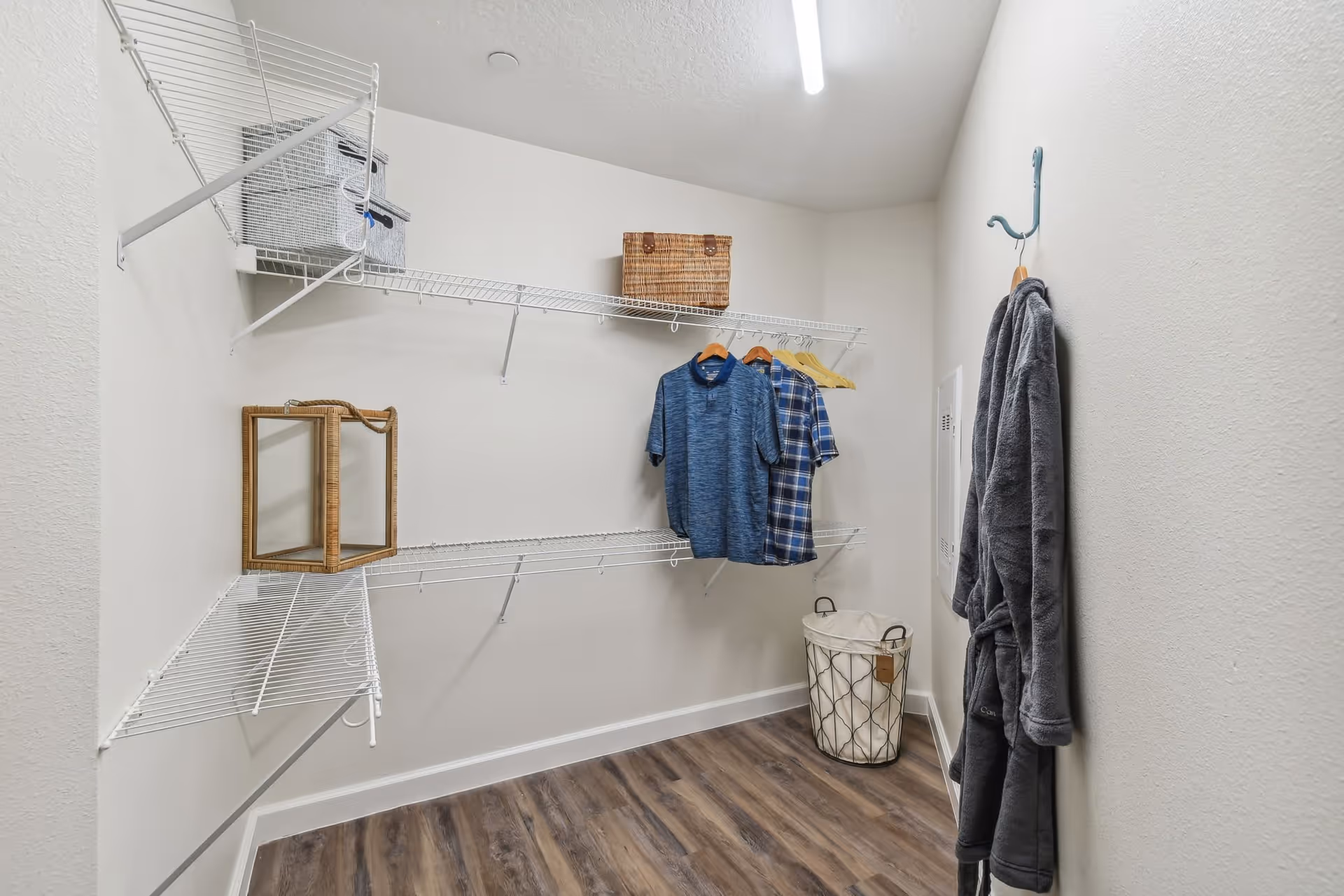 A walk-in closet with white wire shelving on the walls. There are two shirts hanging on wooden hangers, a gray robe hanging on a blue hook, a laundry basket with a metal frame and fabric liner on the floor, and some storage baskets on the shelves. The floor is wood laminate and the walls are painted white.
