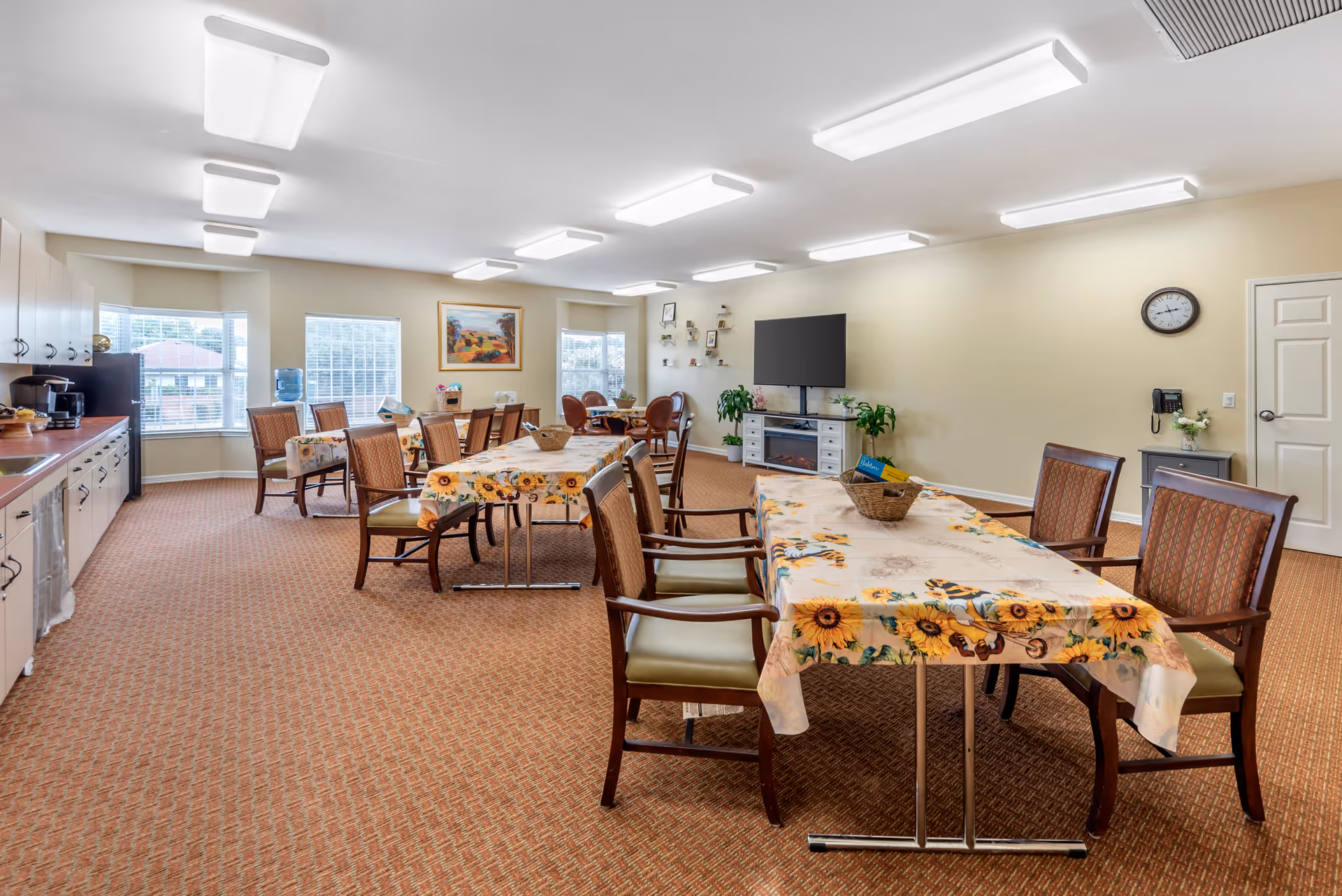 A bright and spacious common dining area with multiple tables covered in sunflower-patterned tablecloths, surrounded by wooden chairs with cushioned seats. The room has beige walls, large windows letting in natural light, a wall-mounted flat-screen TV above a white cabinet with plants on either side, and a clock on the wall. There is a kitchenette area with cabinets, a coffee maker, and a water dispenser along one side of the room.