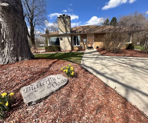 Single-story brick house with a chimney, a front door with a small window, and a large window to the left. A concrete driveway leads up to the entrance. In the foreground, there is a large tree and a rock with the inscription 'Butterfly Oasis' surrounded by mulch and yellow flowers. The sky is blue with some clouds.