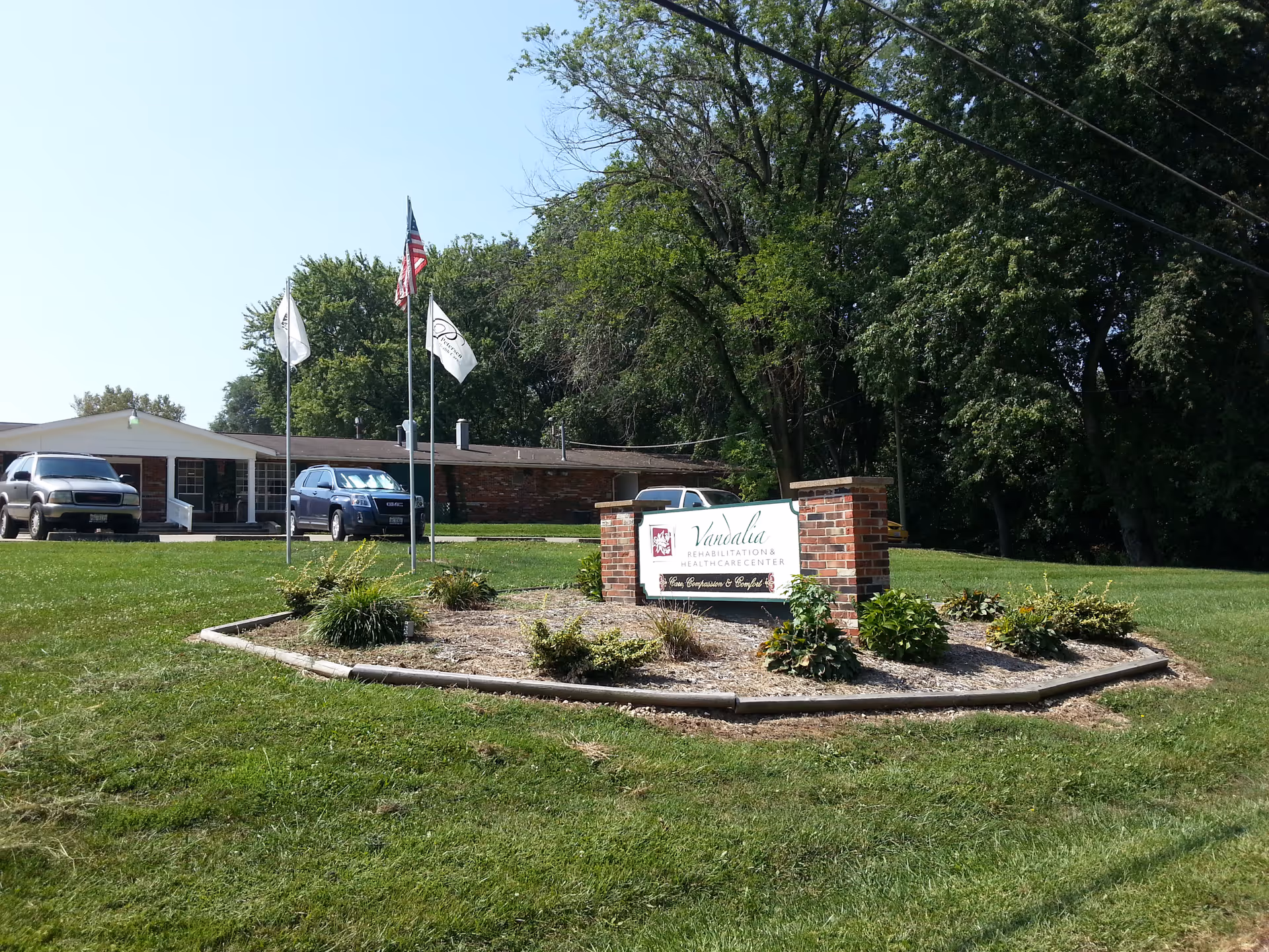 Exterior view of Vandalia Healthcare and Senior Living facility with a brick sign in the foreground surrounded by landscaping. Three flagpoles with flags, two vehicles parked near the building, and trees in the background under a clear sky.