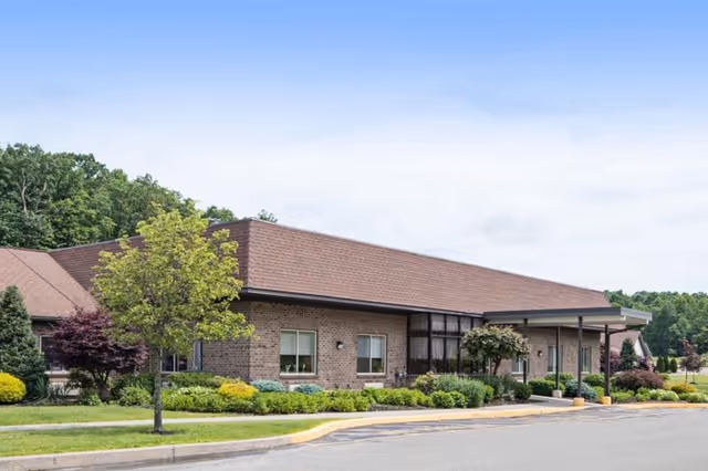 Exterior view of a single-story brick building with a sloped roof, surrounded by well-maintained landscaping including trees, shrubs, and grass under a clear blue sky.