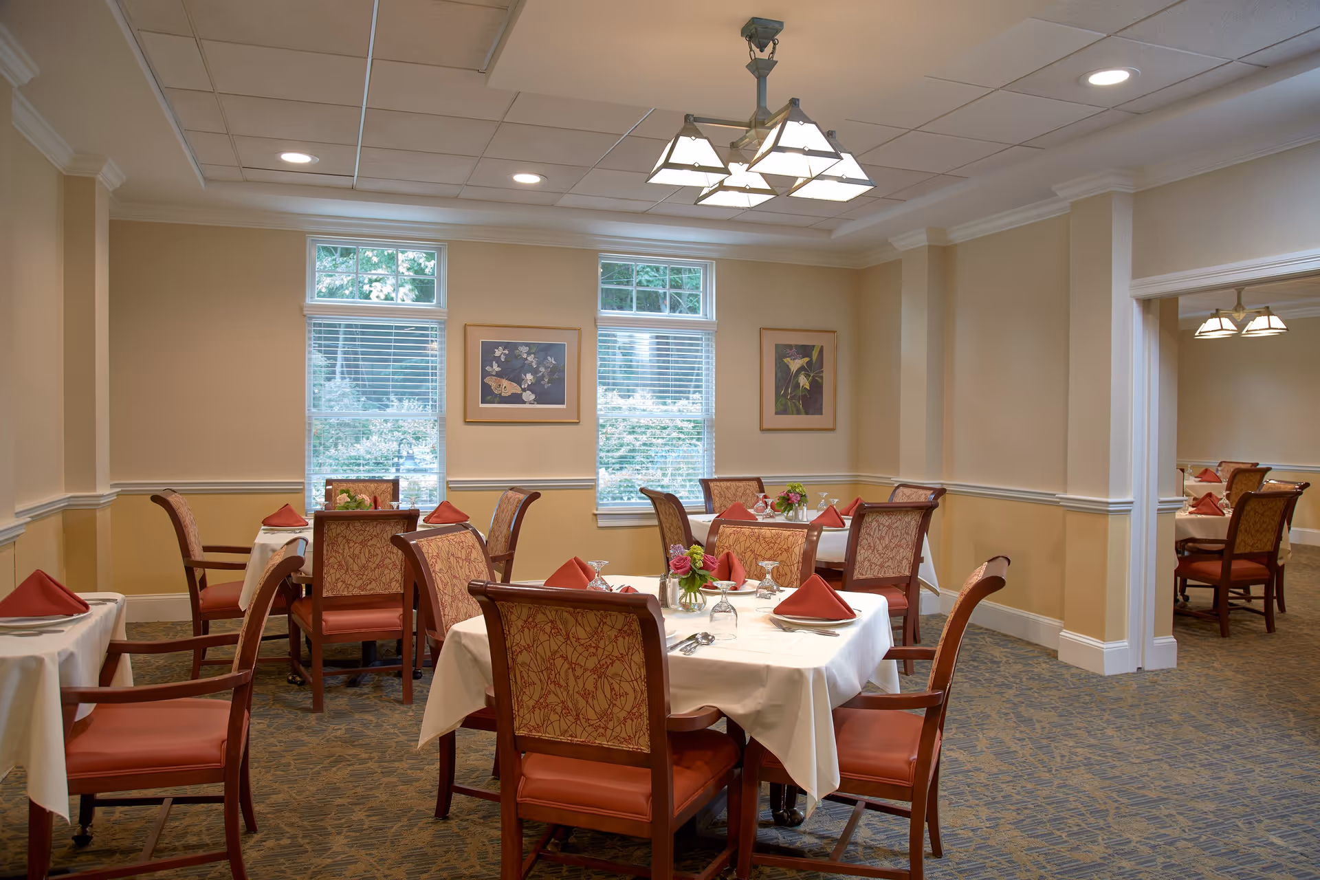 A dining room with several tables covered in white tablecloths, each set with red folded napkins, glassware, and silverware. The room has beige walls, carpeted floors, and windows with white blinds letting in natural light. There are framed floral artworks on the walls and a ceiling light fixture with multiple lamps.