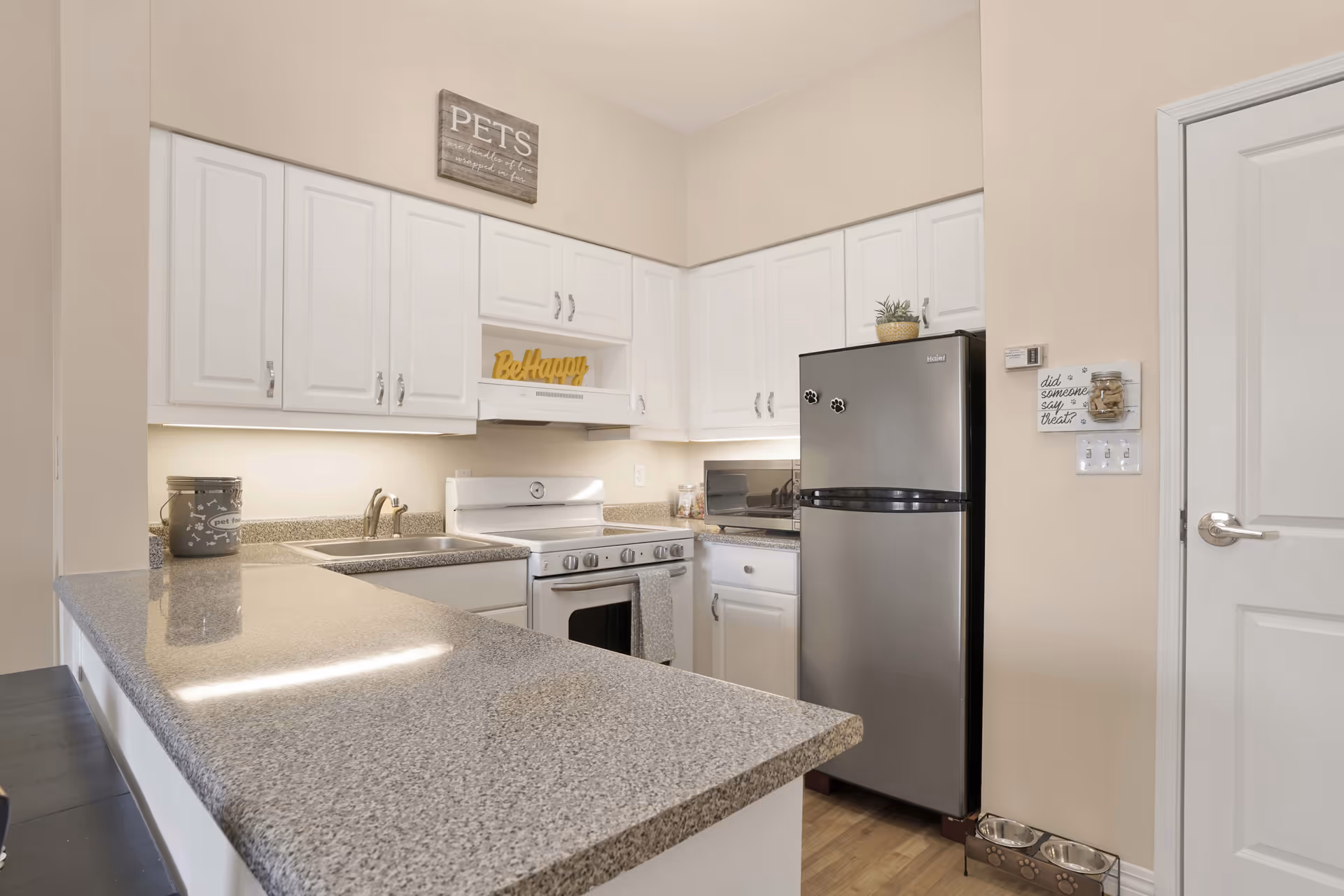 A clean and modern kitchen with white cabinets, a granite countertop, a stainless steel refrigerator, a white stove, and a microwave. There are decorative signs on the walls, including one that says 'PETS' and another that says 'Be Happy'. A pet feeding station with two bowls is on the floor near the refrigerator.