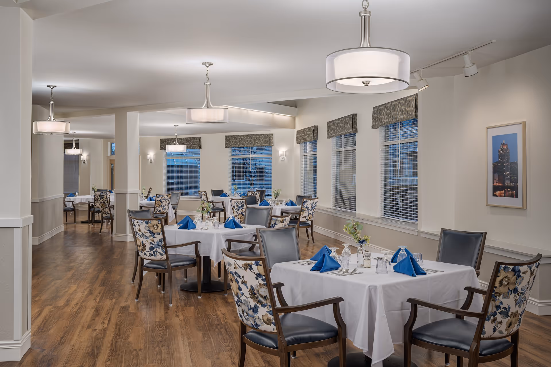 A bright and elegant dining room with multiple tables covered in white tablecloths, each set with blue folded napkins, glassware, and silverware. The chairs have floral patterned backs and dark seats. Large windows with blinds and valances line the walls, and modern pendant lights hang from the ceiling. The floor is wooden, and a framed picture is visible on the wall.