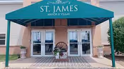 Entrance to St. James Manor & Villas featuring double glass doors under a green awning with the facility name. There are two large planters with greenery on either side of the entrance and a decorative wagon wheel with flowers in the center.