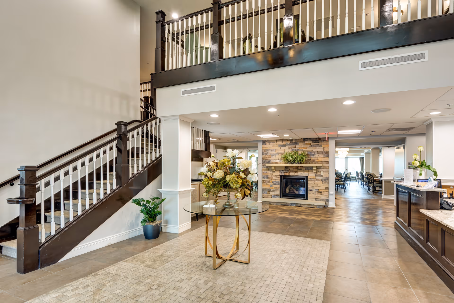 Interior view of a senior living facility lobby area featuring a staircase with dark wood railings and white balusters, a round glass table with a floral arrangement in the center, a stone fireplace with a plant on the mantel, and a reception desk on the right side. In the background, there is a dining area with tables and chairs.