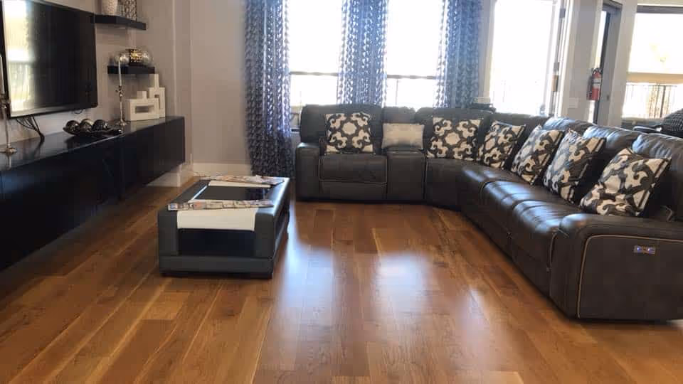 A modern living room with a large dark brown leather sectional sofa adorned with patterned cushions. There is a rectangular coffee table in front of the sofa, and a flat-screen TV mounted on the wall above a floating black media console. The room has wooden flooring and large windows with patterned curtains allowing natural light to enter.