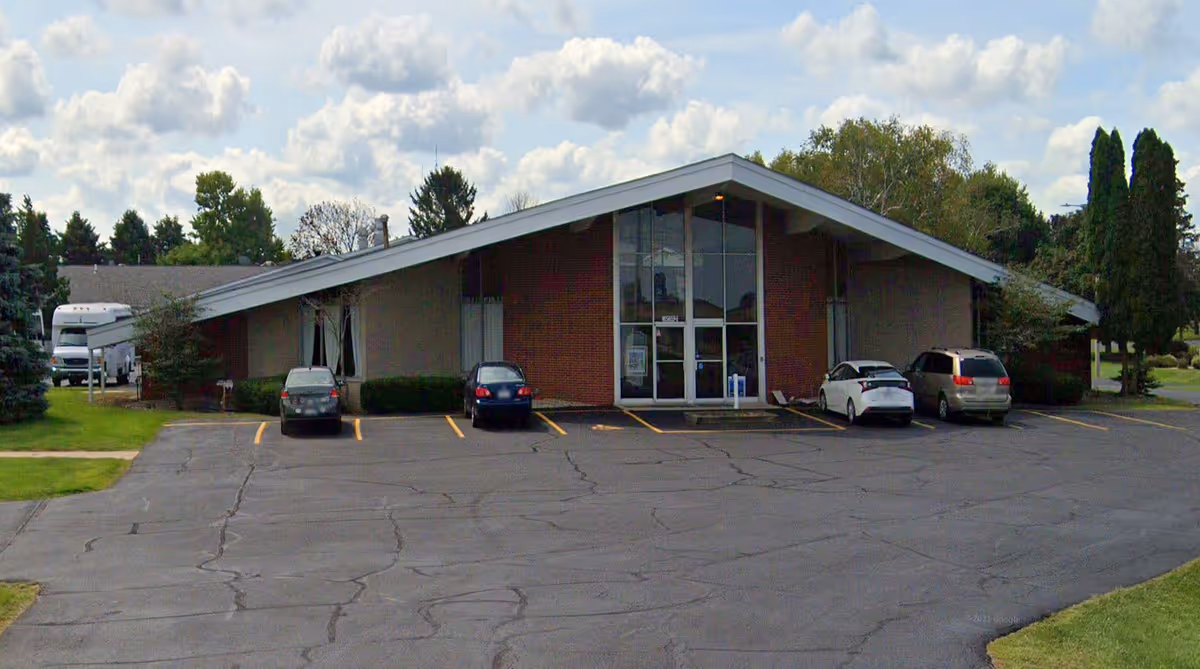 Exterior view of a single-story building with a slanted roof and large glass entrance doors. There are four cars parked in front of the building on an asphalt parking lot. Trees and grass surround the building under a partly cloudy sky.