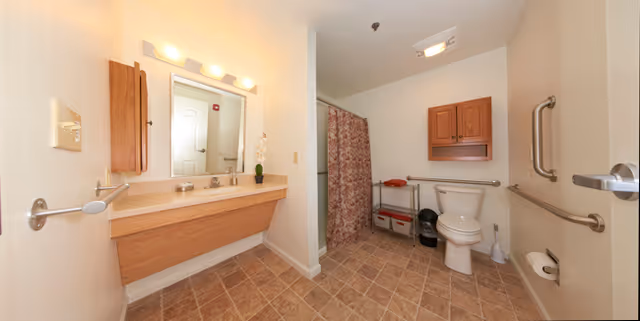A spacious bathroom with beige tiled floor and white walls. There is a wooden vanity with a large mirror and three lights above it on the left side. In the back, there is a shower with a patterned curtain, a white toilet with grab bars on the walls around it, a small metal shelf with towels, and a wooden cabinet mounted above the toilet. The bathroom is equipped with accessibility features including grab bars and a wide layout.