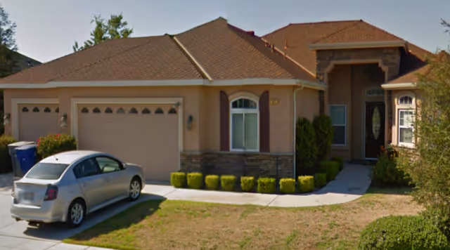 Exterior view of a single-story residential building with a brown roof and beige walls. There is a driveway with a silver car parked on it, a garage with three doors, and a front entrance with a small porch area. The landscaping includes trimmed bushes and a lawn area.