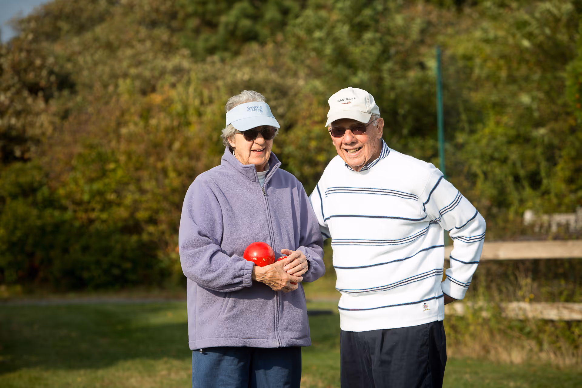Two elderly individuals standing outdoors on a grassy area with trees in the background. One person is wearing a light purple jacket and visor, holding a red ball, while the other is wearing a white and blue striped sweater and a white cap. Both are smiling and wearing sunglasses.