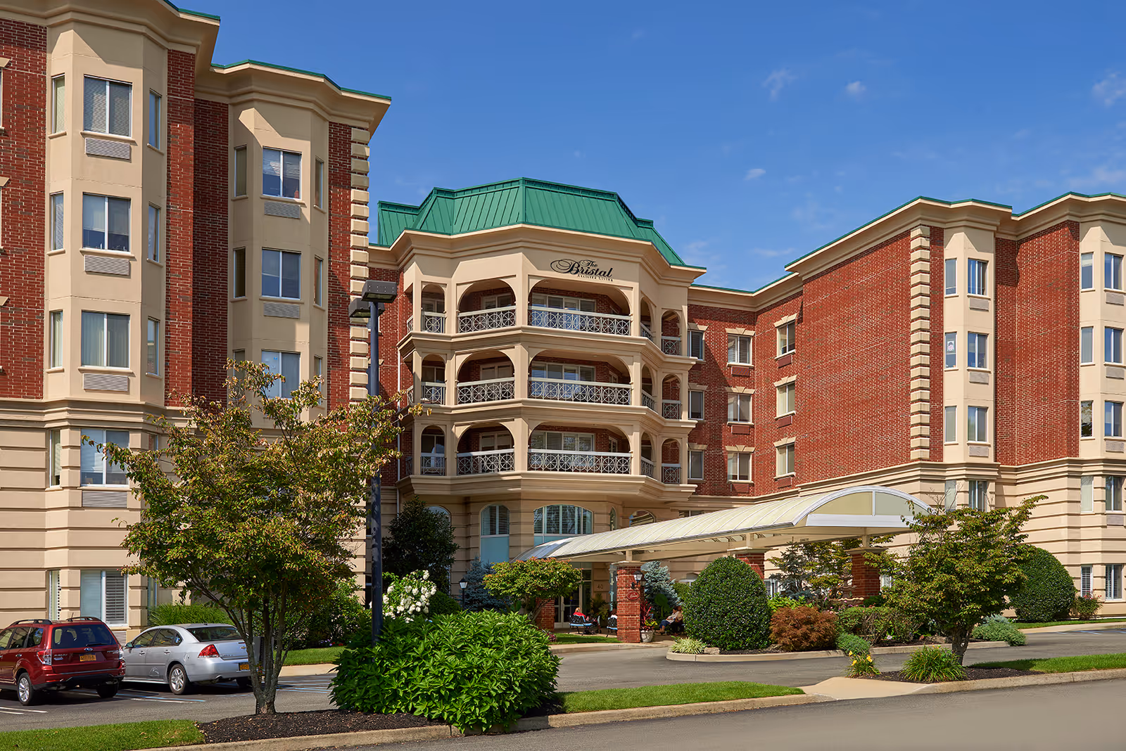 Exterior view of The Bristal Assisted Living at East Meadow building with a red brick and beige facade, green roof accents, balconies, landscaped bushes and trees, and a covered entrance driveway with parked cars nearby under a clear blue sky.
