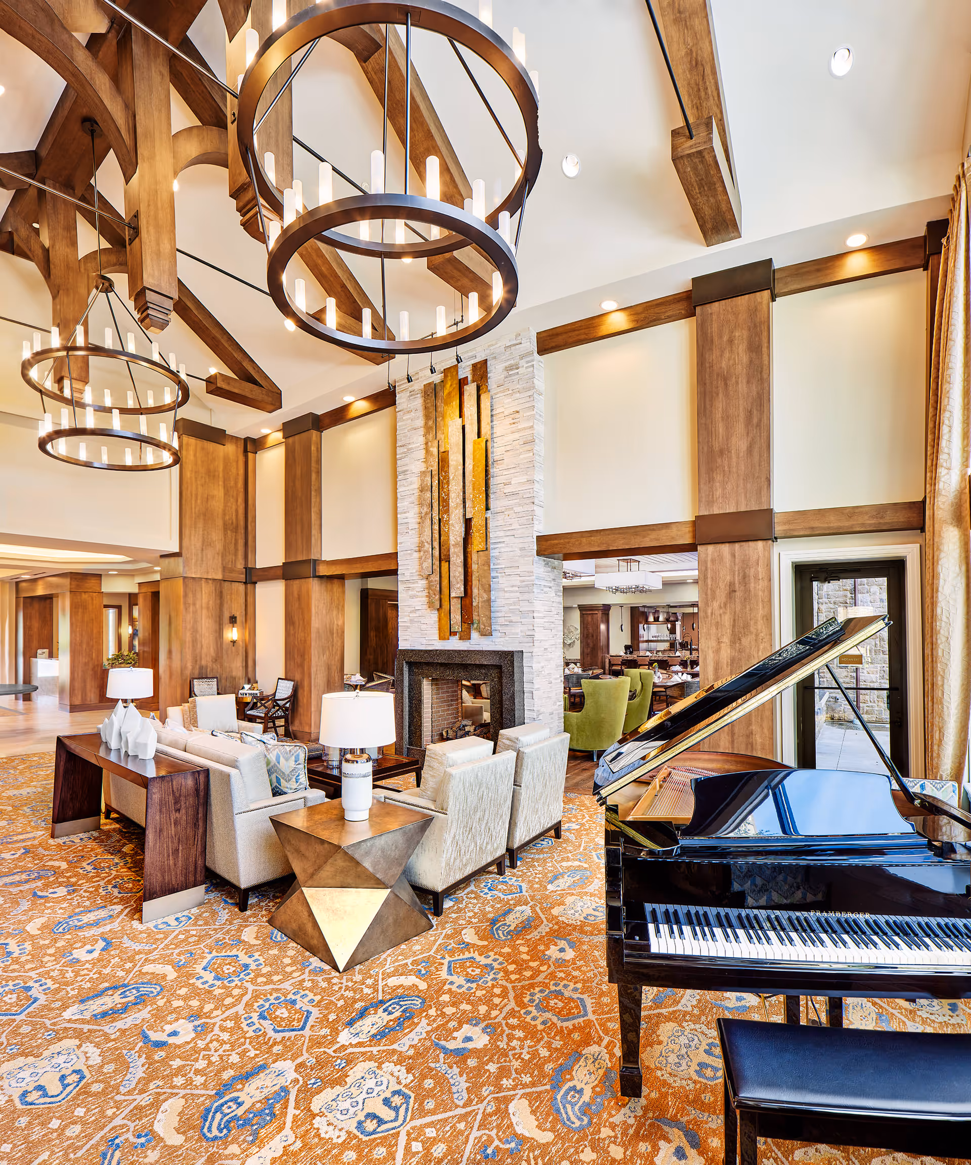 Spacious and elegant living room area with high vaulted ceilings featuring wooden beams and large circular chandeliers. The room includes a grand piano on the right, a modern fireplace with decorative art above it, and comfortable seating arrangements with sofas and chairs on a patterned orange and blue carpet. Large windows with curtains allow natural light to enter the space.