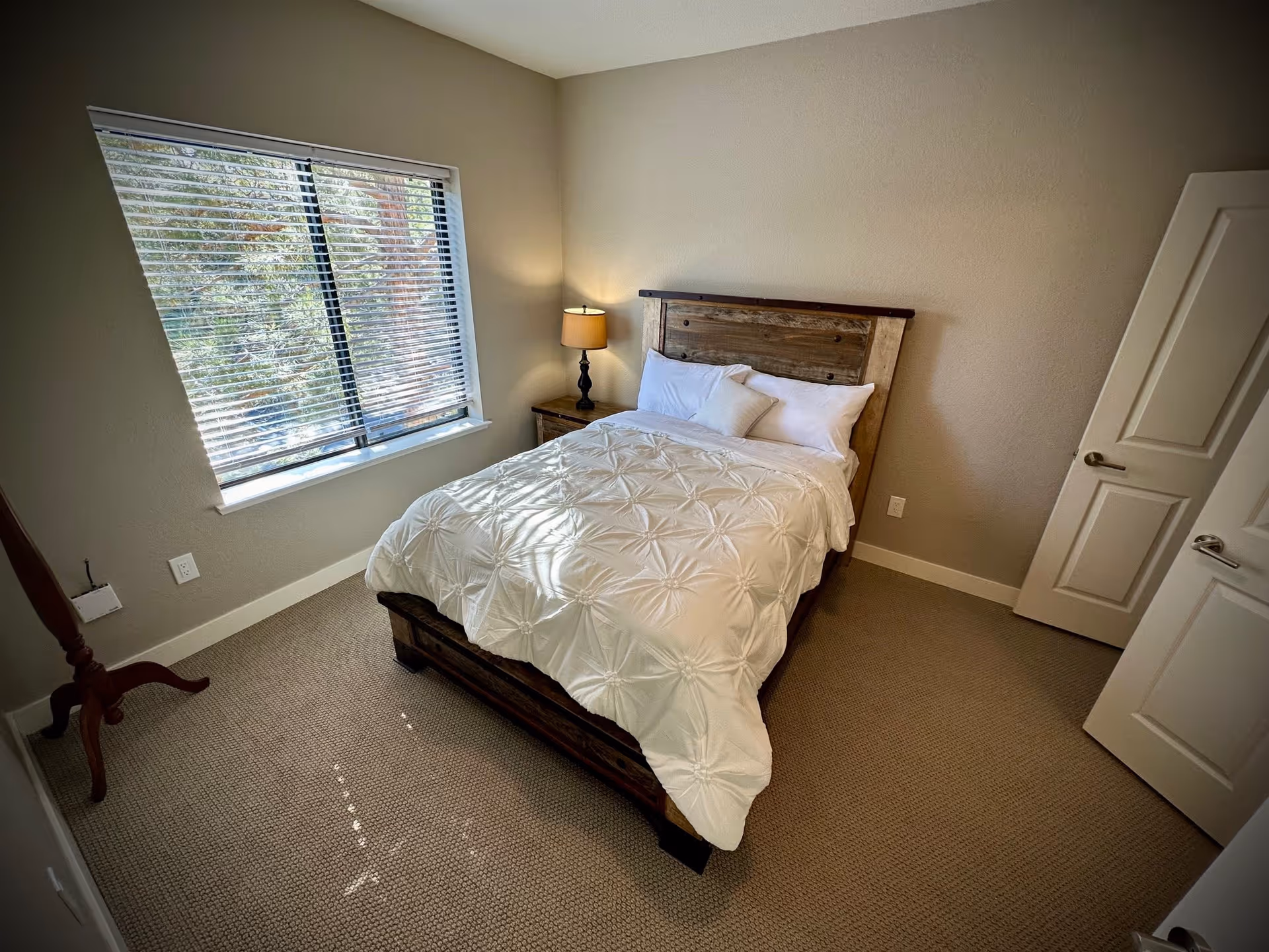 A small bedroom with a wooden bed frame and a white comforter. There are three pillows on the bed. A wooden nightstand with a lamp is positioned next to the bed. The room has beige walls and carpeted floor. A window with blinds lets in natural light, and a partially open white door is visible.