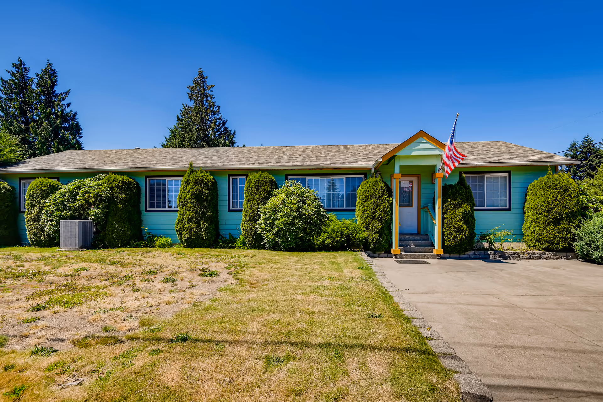 Single-story turquoise building with a gray shingled roof, surrounded by neatly trimmed bushes and trees. There is a small covered porch with an American flag mounted on a pole near the entrance. The lawn in front is patchy with some dry grass, and a concrete driveway leads up to the porch. The sky is clear and blue.