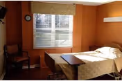 A small, simple bedroom with a single bed covered in a beige quilt, a wooden chair with armrests, a wall-mounted television, a clock on the wall, and a window with blinds and a beige valance letting in natural light.