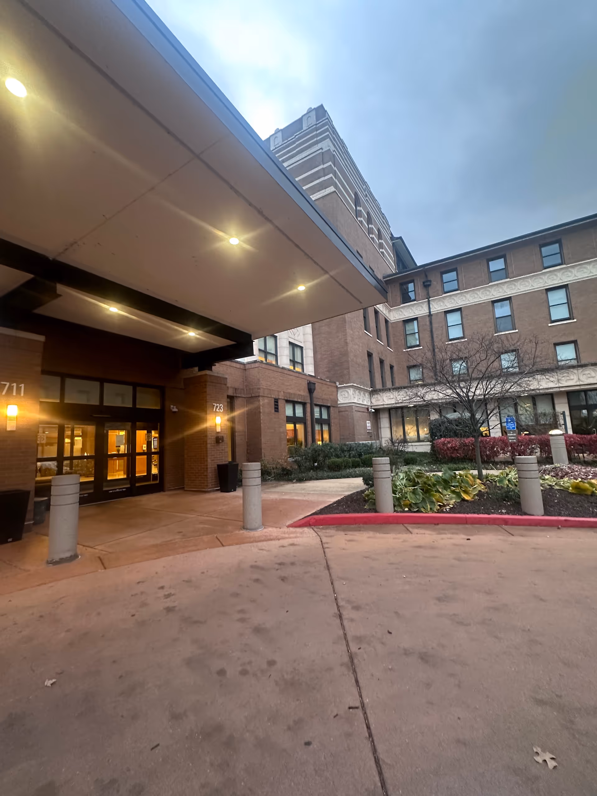 Entrance area of a multi-story brick building with a covered drop-off zone, illuminated lights under the canopy, and landscaped garden beds with plants and a small tree near the sidewalk.