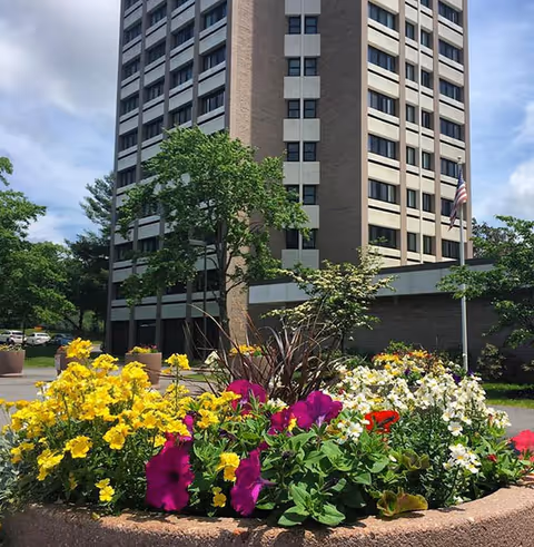A tall multi-story building with a flower bed in the foreground containing yellow, purple, white, and red flowers. Trees and an American flag on a flagpole are visible near the building under a partly cloudy sky.