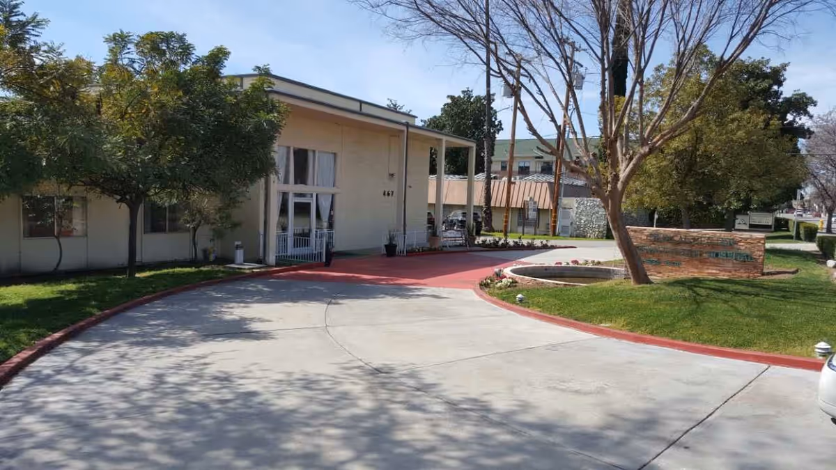 Exterior view of a single-story building with a curved driveway and landscaped greenery including trees and grass. There is a sign on the right side near the driveway entrance, and the building has large windows and a covered entrance.