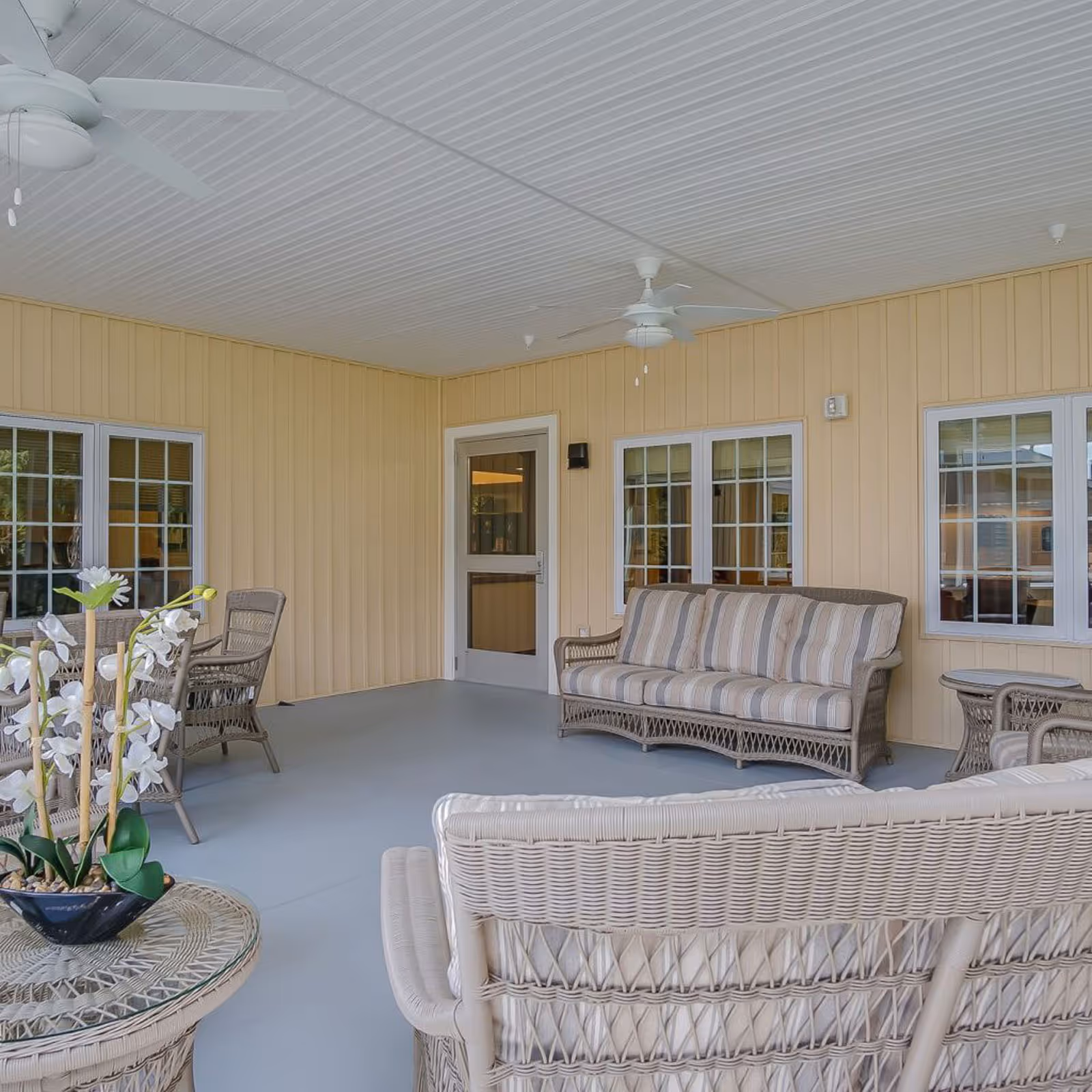 Covered outdoor patio area with beige walls and ceiling fans. The space is furnished with wicker furniture including a striped cushioned sofa, chairs, a round table with a flower arrangement, and a small side table. Multiple windows and a door lead inside the building.