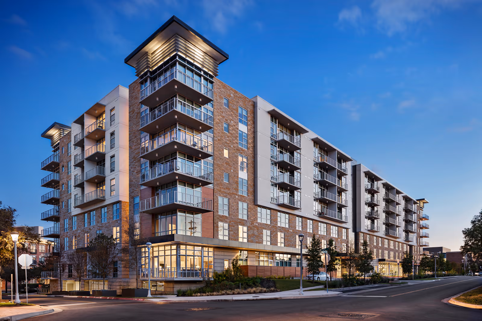 Exterior view of a modern multi-story residential building with balconies and large windows during dusk, with street lamps and trees along the sidewalk.