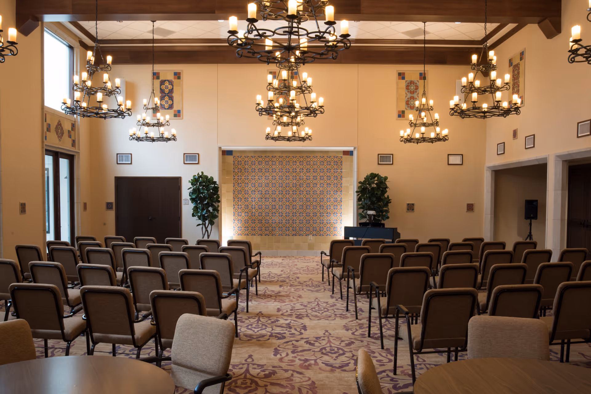 A large room set up with rows of brown cushioned chairs facing a tiled wall with intricate patterns. The room features multiple ornate chandeliers hanging from a high ceiling with wooden beams. There are two potted plants on either side of the tiled wall and round tables with chairs in the foreground. The walls are decorated with small framed artworks and tiles.