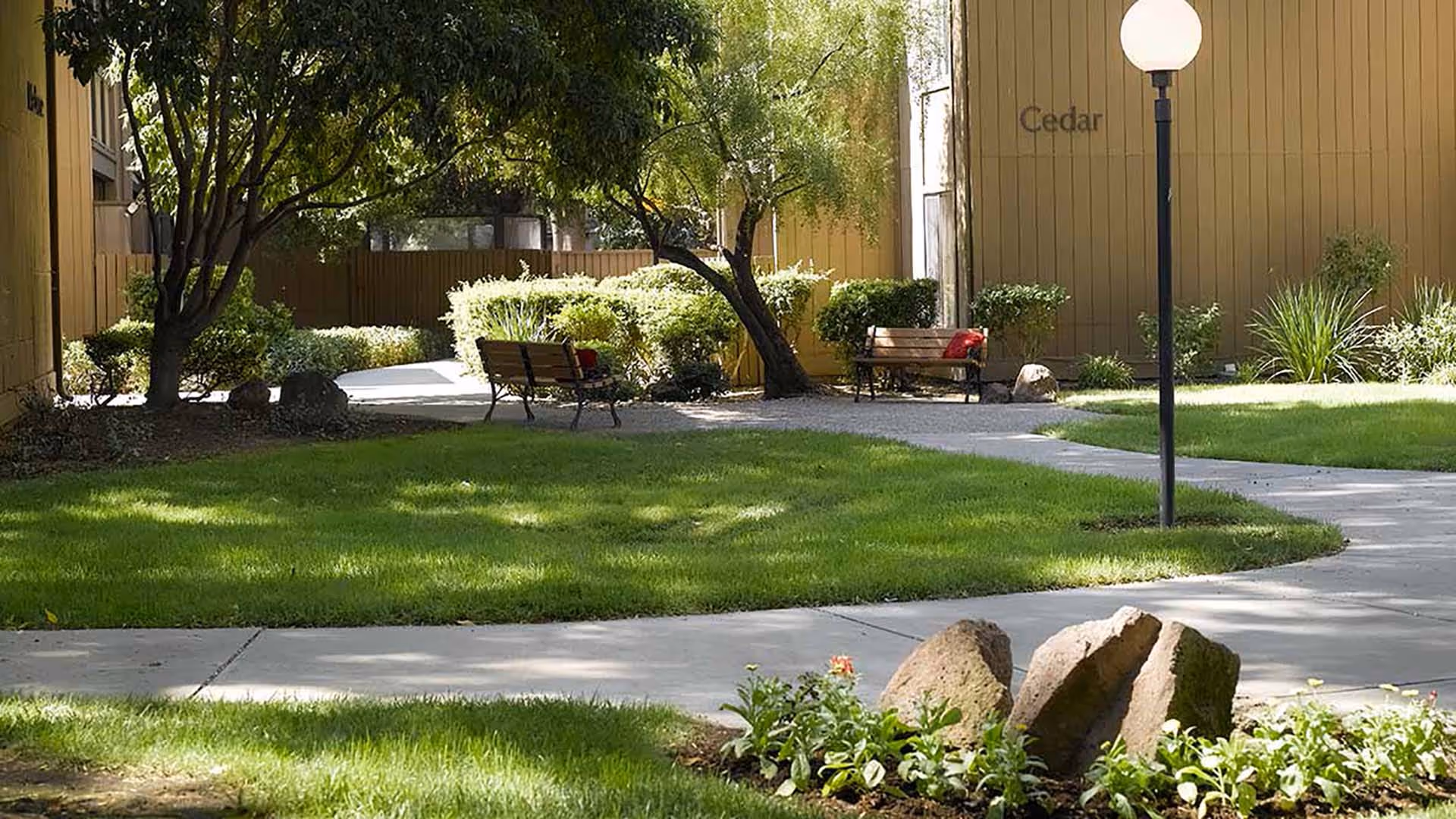 A peaceful outdoor garden area with green grass, trees, bushes, and a paved walkway. Two wooden benches with red cushions are placed near the trees. A lamppost with a round white light stands near the walkway. The building wall in the background has the word 'Cedar' written on it.