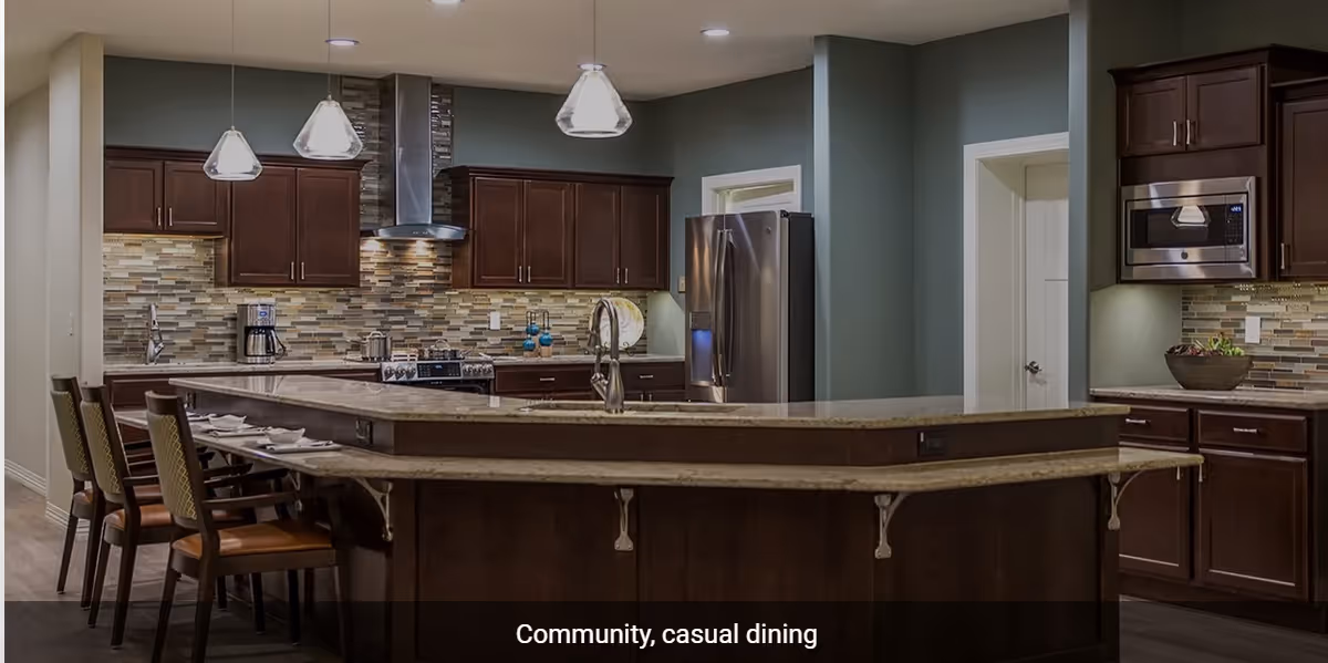 A modern kitchen area with dark wood cabinets, a large granite countertop island with seating, stainless steel appliances including a refrigerator, stove, and microwave, and pendant lights hanging from the ceiling. The backsplash features a mosaic tile design.