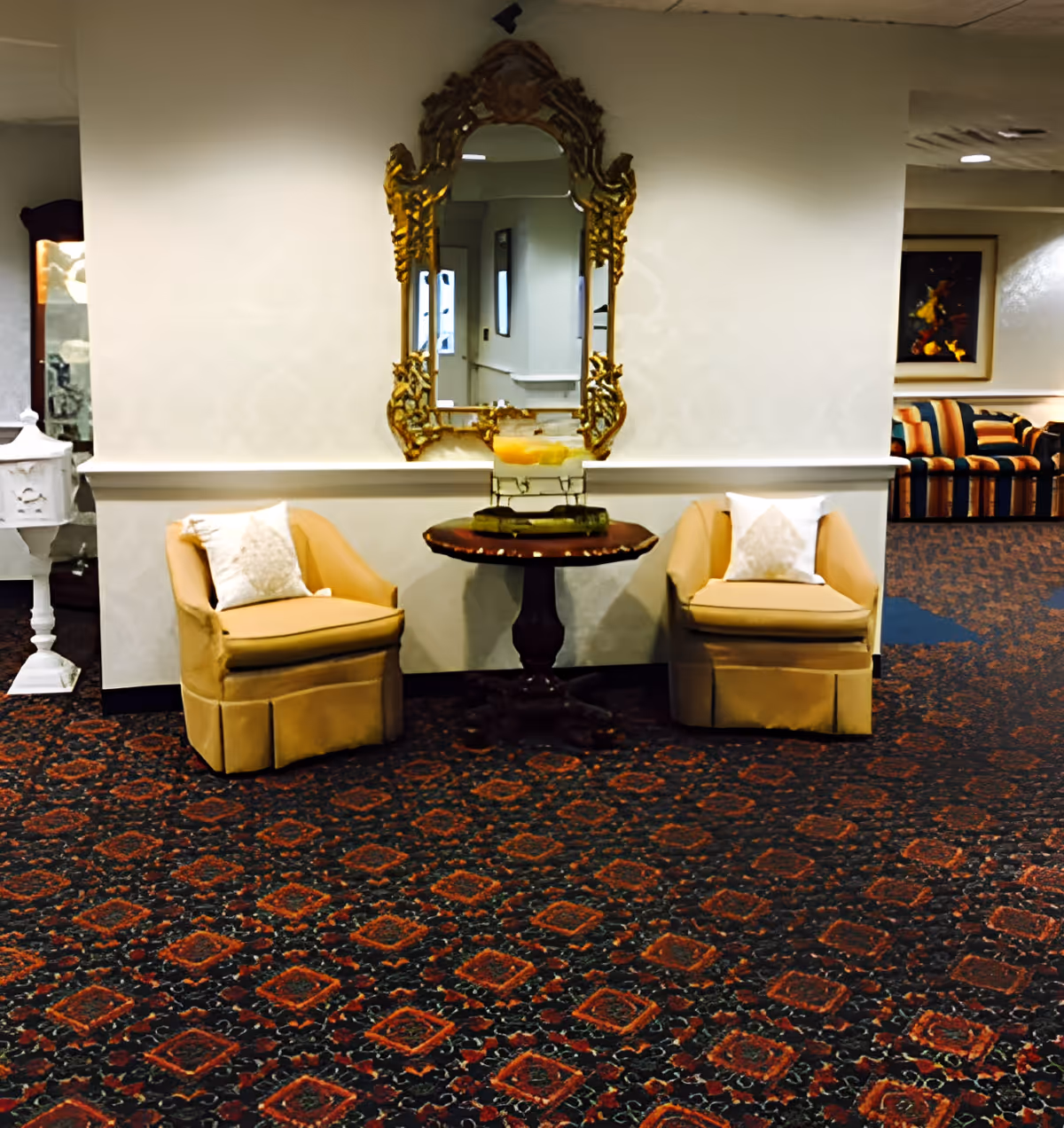 A cozy seating area with two beige armchairs each with a white pillow, positioned on either side of a small round wooden table. Above the table is an ornate gold-framed mirror reflecting part of the room. The floor is covered with a patterned carpet in dark red and black tones. In the background, there is a striped sofa and a framed painting on the wall.