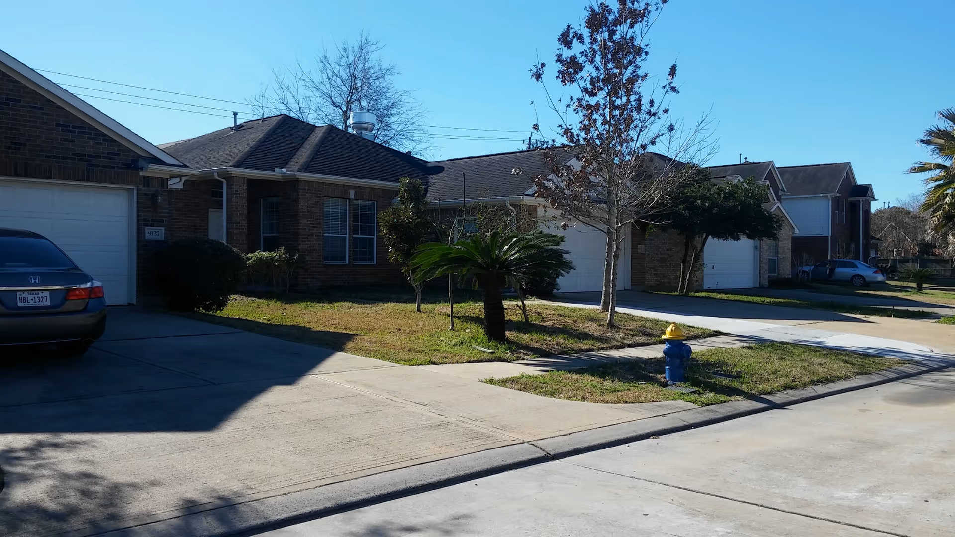 A residential street view showing single-story brick houses with garages, driveways, small front lawns with trees and shrubs, a blue fire hydrant with a yellow top, and parked cars under a clear blue sky.