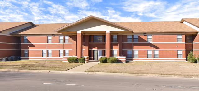 Front exterior view of a two-story brick building with a peaked roof and a covered entrance supported by four columns. The building has multiple windows and is surrounded by a small lawn and a sidewalk leading to the entrance. The sky is partly cloudy.