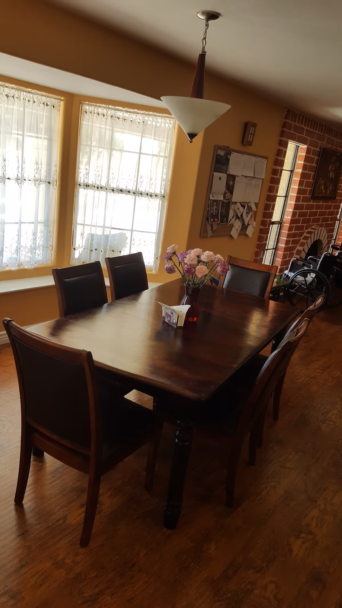 A dining room with a wooden table surrounded by six chairs. A vase with pink and purple flowers is placed in the center of the table. The room has large windows with lace curtains allowing natural light to enter. There is a brick fireplace and a bulletin board on the wall in the background.