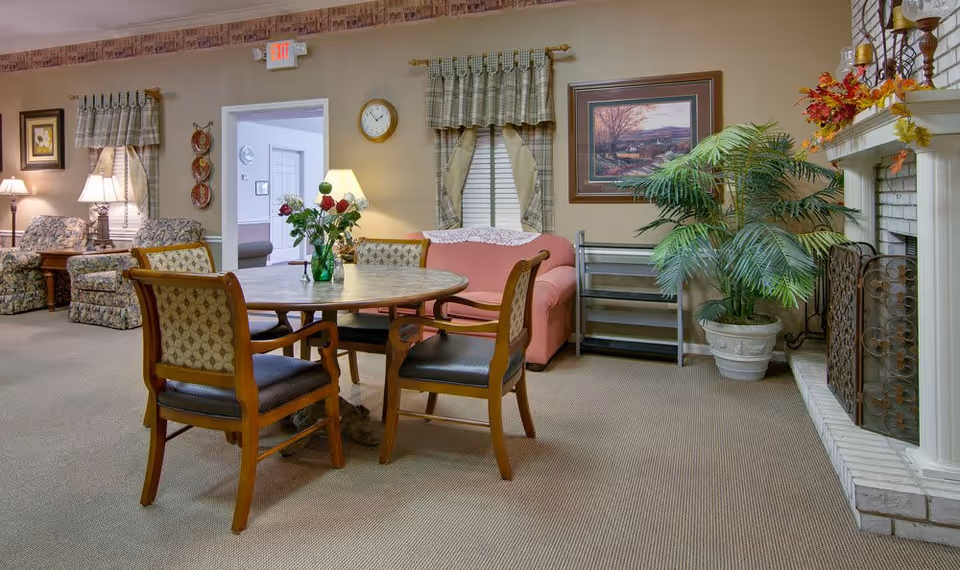 A cozy living room area with a round wooden table surrounded by four chairs, a pink loveseat with a lace doily on the back, a large potted plant, and a white brick fireplace decorated with autumn leaves. The room has beige walls with framed artwork, a clock, and windows with plaid curtains. There are also two floral armchairs and lamps in the background.