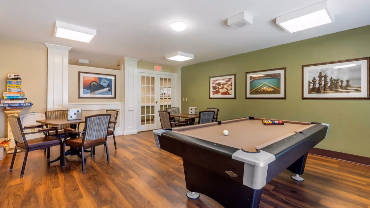 A recreational room with a pool table in the foreground and several tables with chairs in the background. The walls are painted green and decorated with framed pictures of billiards and chess. There is a stack of board games on a small table near the left wall, and the floor is wooden. The room is well-lit with ceiling lights and has double glass doors at the back.