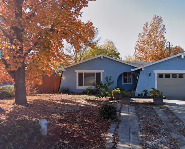 Single-story blue house with white trim surrounded by autumn trees with orange and yellow leaves. The front yard is covered with fallen leaves and has a concrete driveway leading to a garage on the right side.