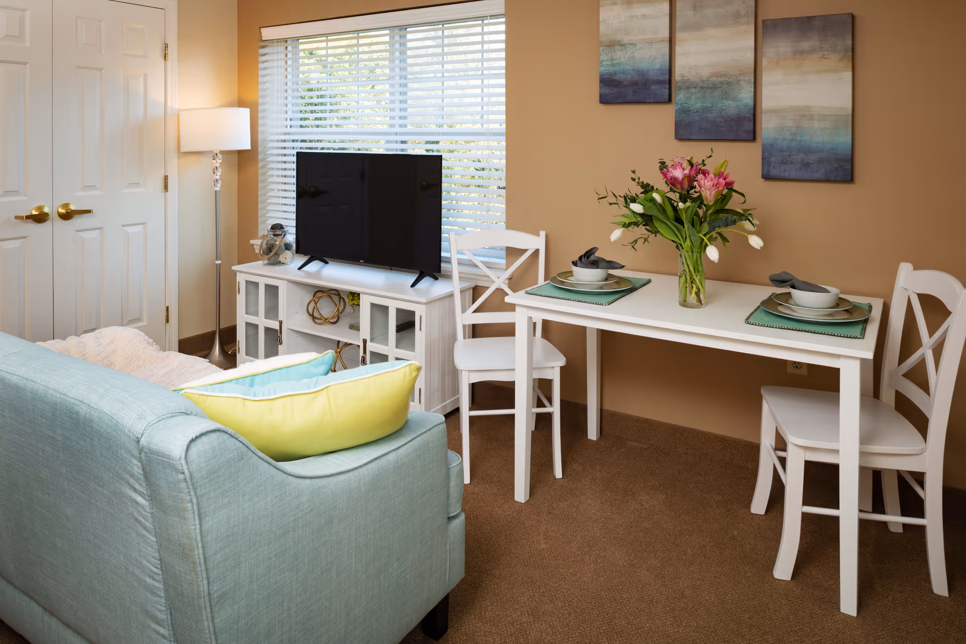 A cozy living and dining area with a light blue sofa adorned with yellow and blue pillows, a white dining table set for two with plates, bowls, and napkins, and a vase of fresh flowers. A TV sits on a white cabinet in front of a window with blinds, and three abstract paintings hang on the beige wall.