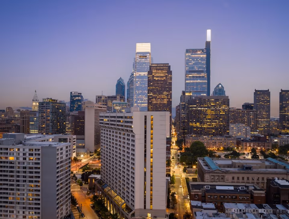 A cityscape view of downtown Philadelphia at dusk, featuring tall buildings with lights on, including the Comcast Center and Liberty Place towers, with streets and smaller buildings in the foreground.