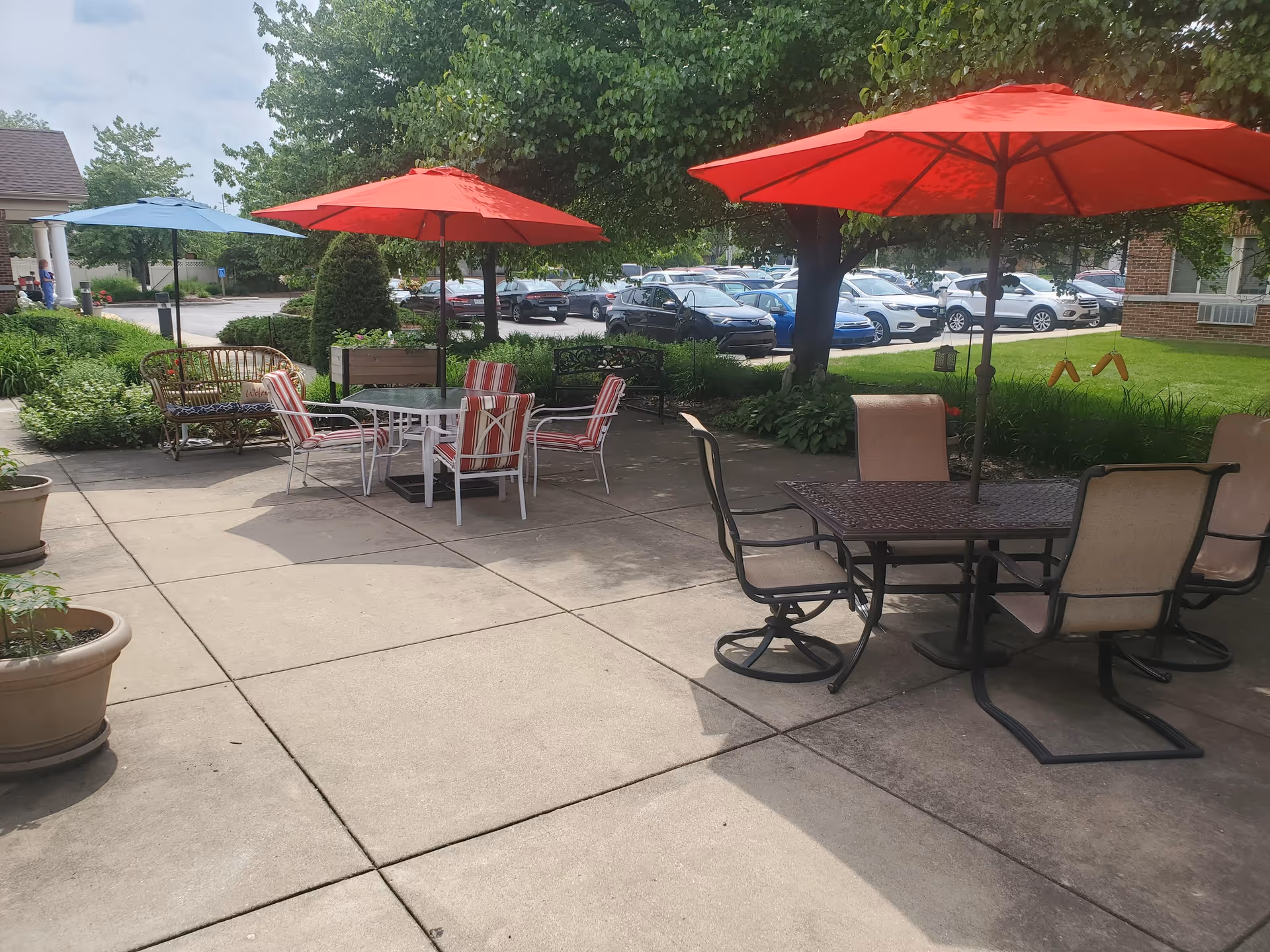 Outdoor patio area with several tables and chairs under large umbrellas, surrounded by greenery and trees. There are parked cars visible in the background and a building partially visible on the right side.