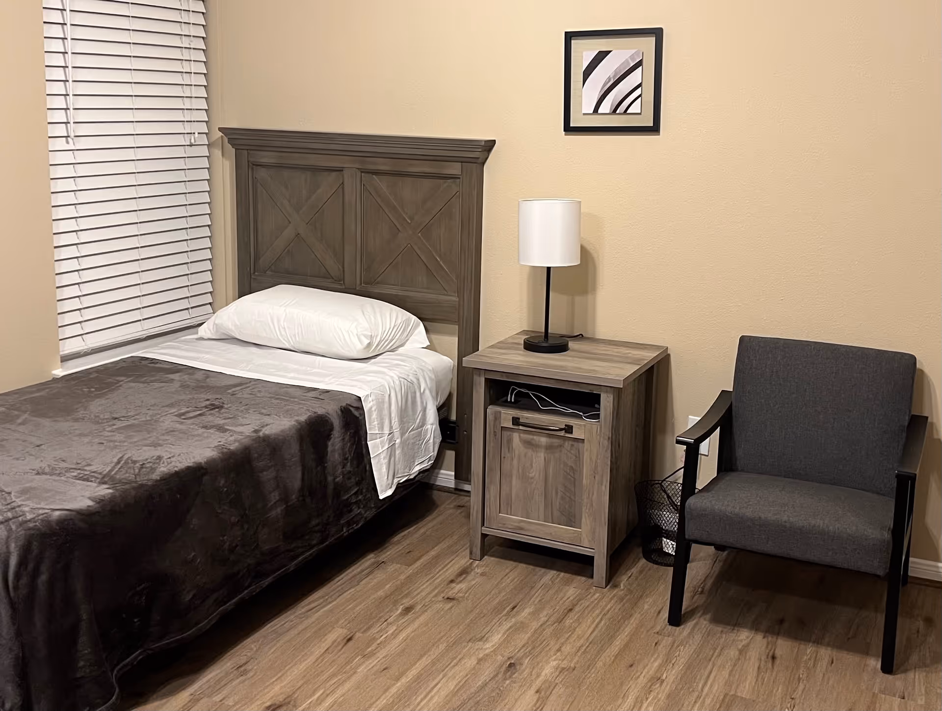 A small bedroom with a single bed featuring a wooden headboard, a white pillow, white sheets, and a dark gray blanket. Next to the bed is a wooden nightstand with a white lamp on top. To the right of the nightstand is a gray upholstered armchair with black armrests. The room has light beige walls, a window with white blinds, and wood flooring. A small framed abstract artwork hangs on the wall above the nightstand.