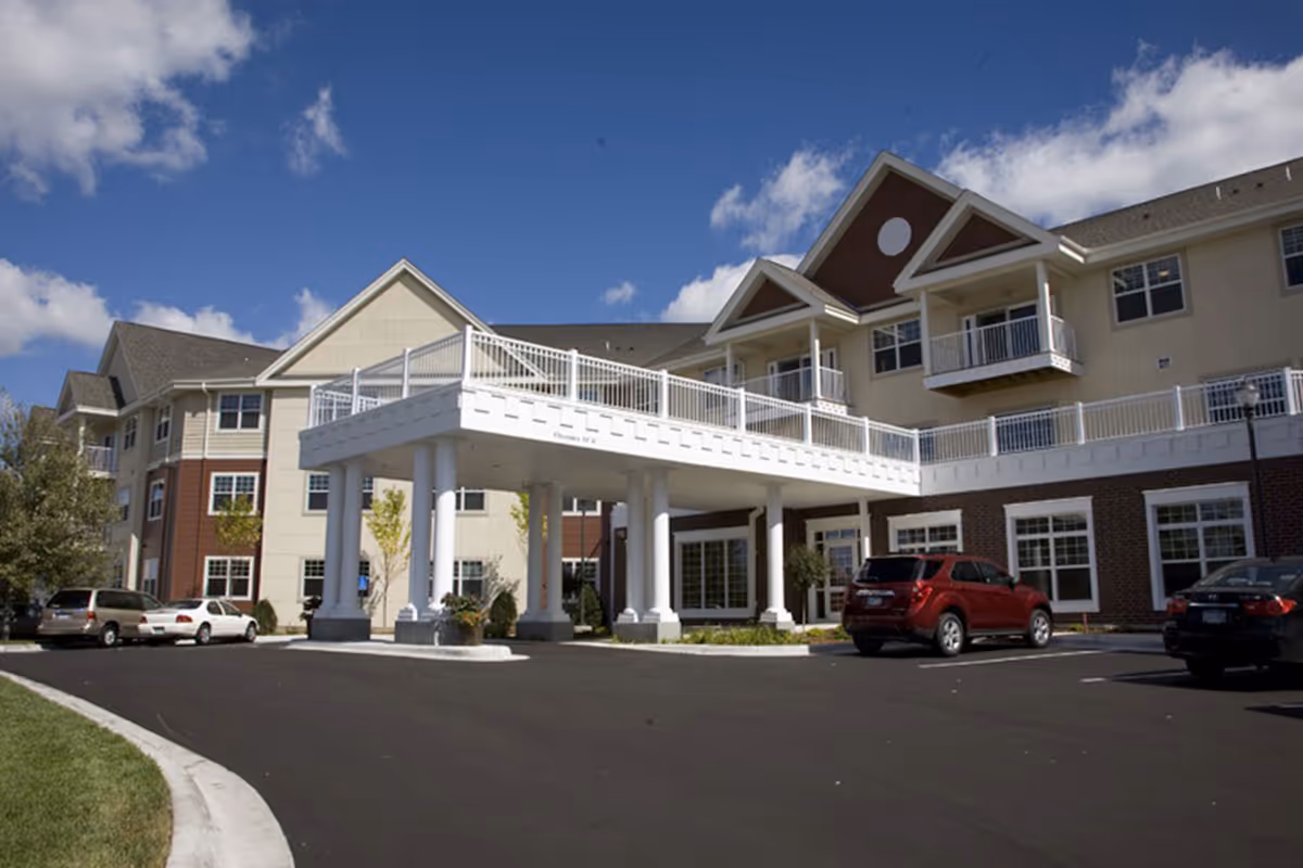 Exterior view of Cherrywood Pointe Assisted living facility showing a multi-story building with a covered entrance supported by white columns, several parked cars, and a clear blue sky with some clouds.
