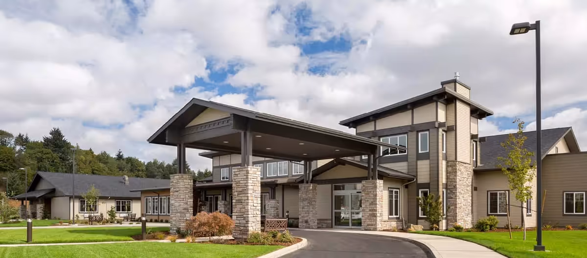 Exterior view of a modern senior living facility building with a covered entrance supported by stone pillars, surrounded by green lawns and trees under a partly cloudy sky.