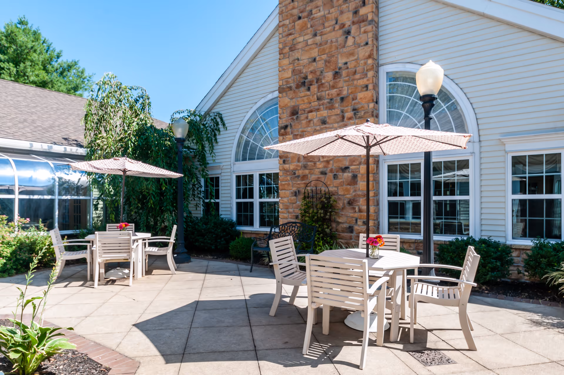 Outdoor patio area with two tables, each surrounded by four chairs and shaded by large umbrellas. The patio is adjacent to a building with large arched windows and a stone chimney. There are lamp posts and greenery around the patio under a clear blue sky.